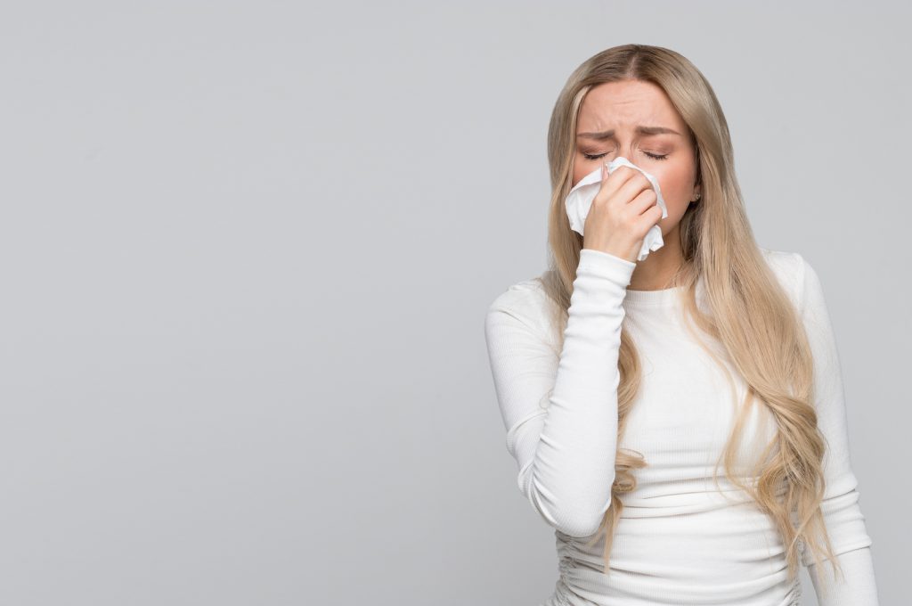 Beautiful unhealthy Caucasian female with paper napkin sneezing, experiences allergy symptoms, caught a cold, closed eyes.Sick desperate woman has flu. Rhinitis, cold, sickness, allergy concept