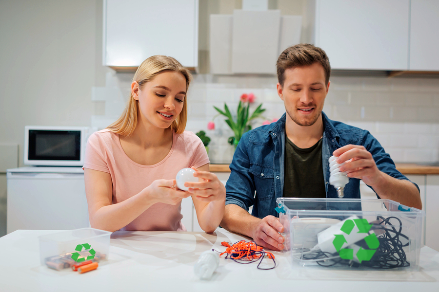 Recycling, reuse, energy. Young family sorting light bulbs, batteries, other electronic waste into containers with recycling symbol while sitting at kitchen
