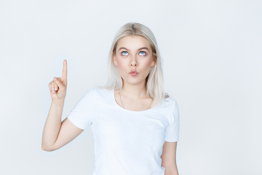 Funny young woman in white shirt pointing finger up isolated over grey background. teen blond girl making grimace and looking up