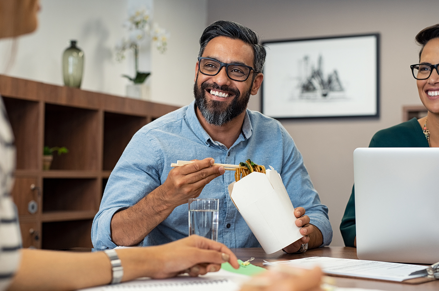 Multiethnic business man with colleagues having chinese take away food. Friendly businessman and casual businesswoman eating noodles. Mature latin man eating lunch meal while talking to creative team.