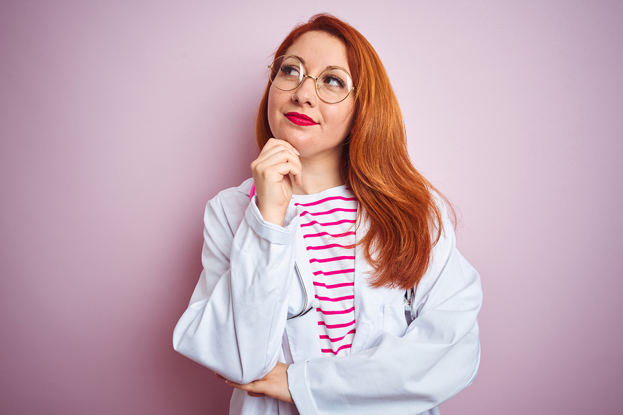 Young redhead doctor woman wearing glasses over pink isolated background with hand on chin thinking about question, pensive expression. Smiling with thoughtful face. Doubt concept.
