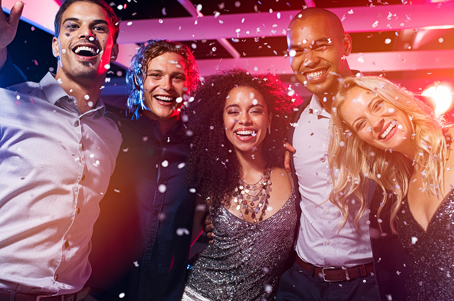 Happy young friends dancing together at party night with confetti. Group of beautiful women and elegant men and looking at camera while partying in nightclub. Group of smiling multiethnic people.