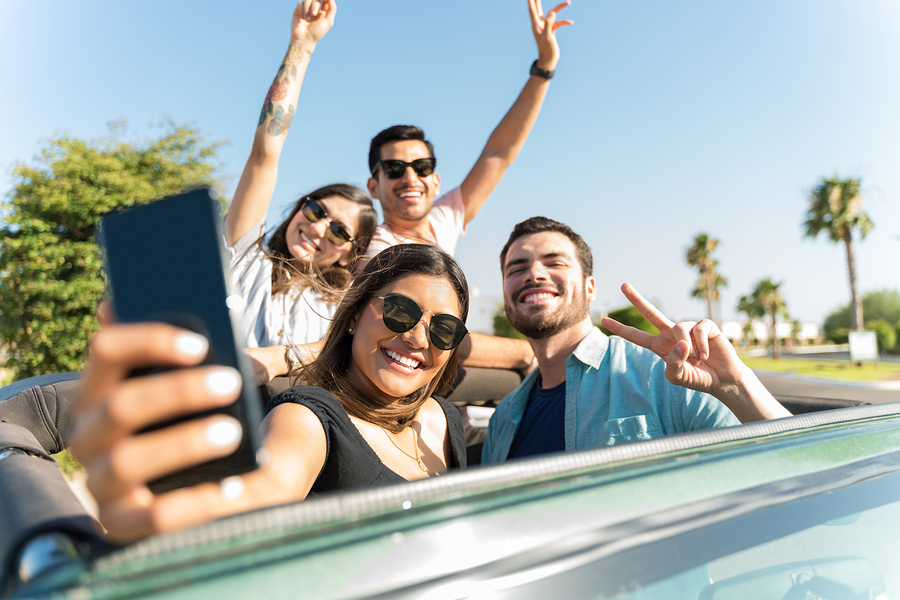 Hispanic woman making memories with friends on smartphone in SUV during roadtrip