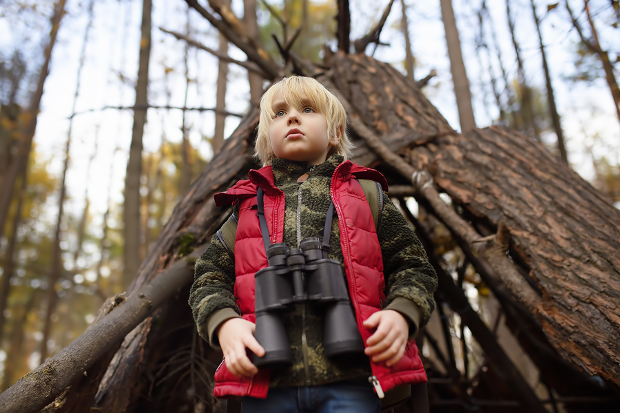 Little boy scout with binoculars during hiking in autumn forest. Behind the child is teepee hut. Concepts of adventure, scouting and hiking tourism for kids.