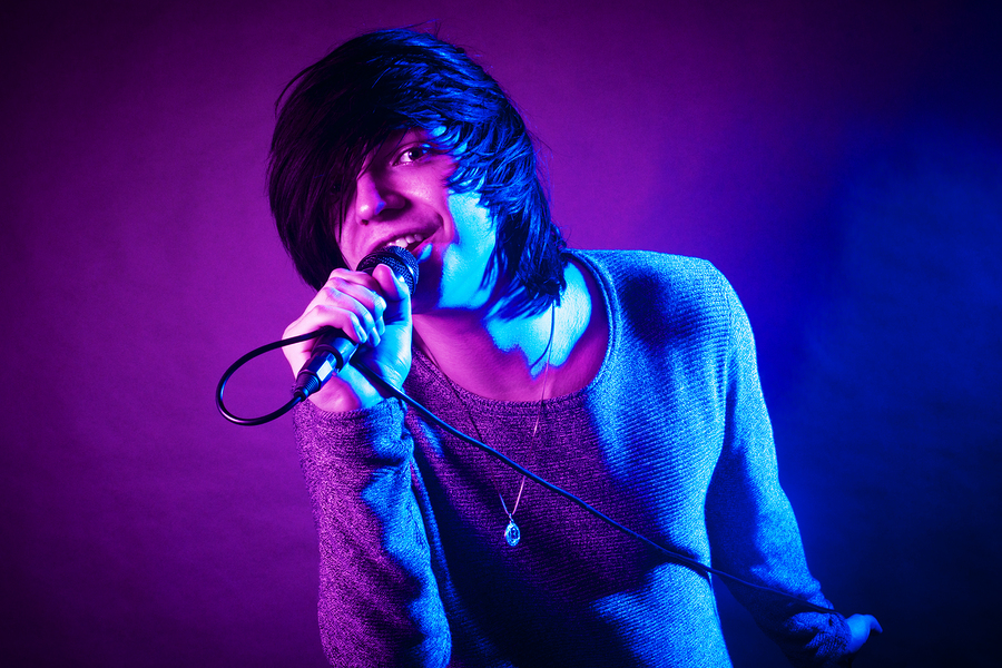 The handsome young emo guy is singing in microphone on purple and blue concert lighting.