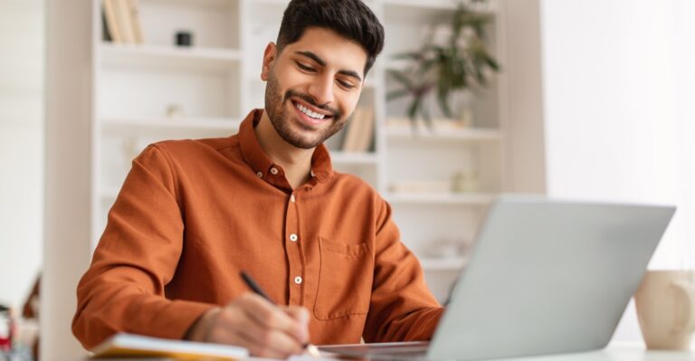 Smiling,Arab,Man,Using,Laptop,Working,With,Documents,At,Home